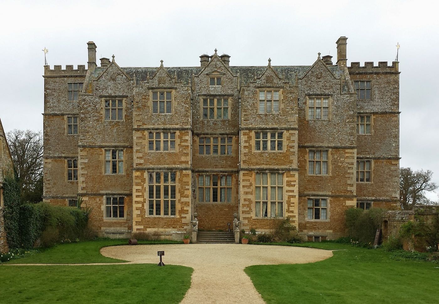 The tall stone front of Chastleton House, with rows of mullioned windows and a gravel path across the lawn to the door
