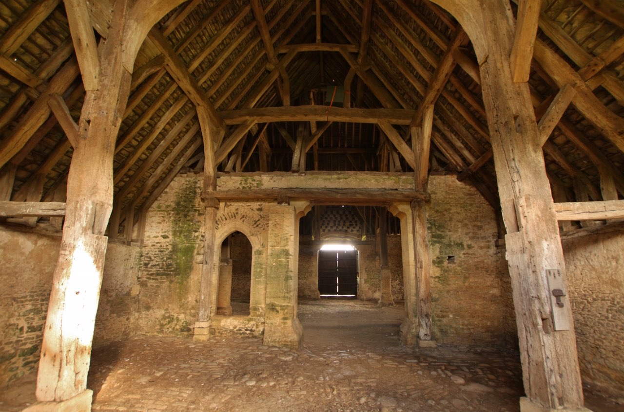 Inside a medieval barn: massive timber posts and braces under a high, dark roof, with daylight coming only through a doorway at the far end