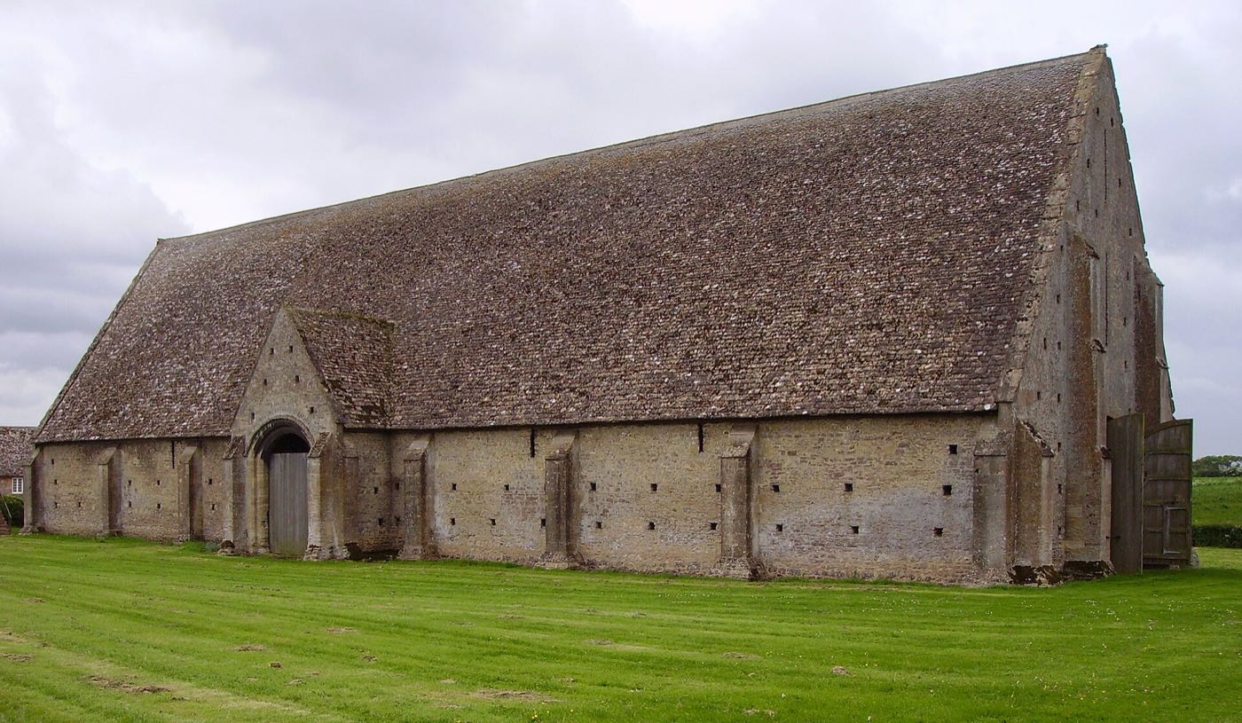 A long medieval stone barn with a steep stone-tiled roof and a gabled porch, standing on a lawn at Great Coxwell